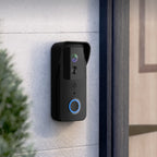 A black smart video doorbell mounted on a light gray concrete exterior wall next to a dark wooden door frame, with a green plant in the foreground.