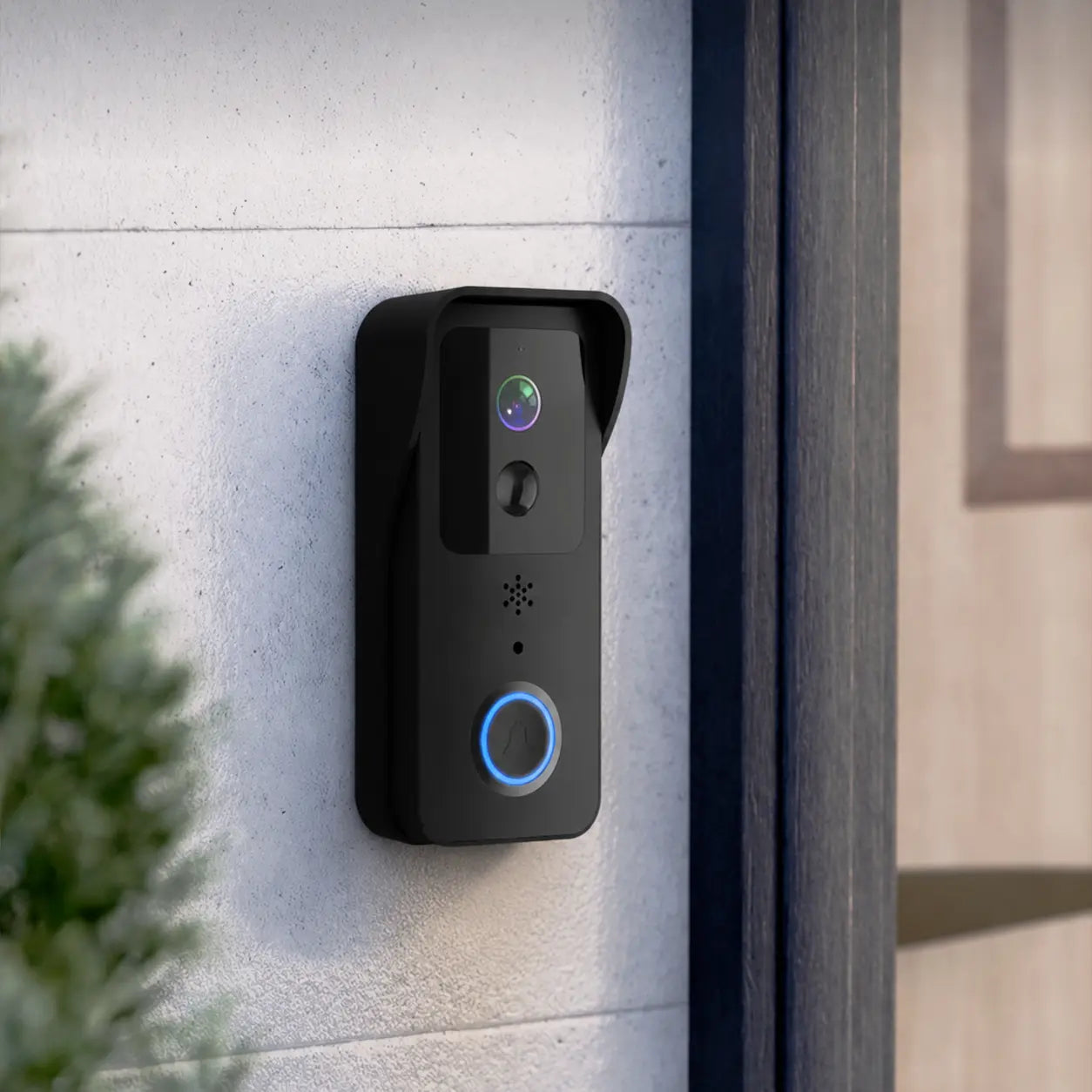 A black smart video doorbell mounted on a light gray concrete exterior wall next to a dark wooden door frame, with a green plant in the foreground.