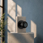 A matte black smart doorbell with a circular touch screen is mounted on a light gray concrete block against a textured blue-gray wall with soft window shadows.