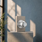 A modern chrome shower valve trim with a round control knob mounted on a square concrete display block against a textured blue-gray wall with soft window shadows.