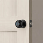 A modern black smart doorbell with a blue ring indicator light mounted on a light wood paneled door next to a metal latch.