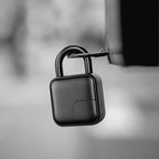 A black square-shaped electronic lock or padlock with a curved shackle, shown hanging from a metal bracket against a blurred gray background.