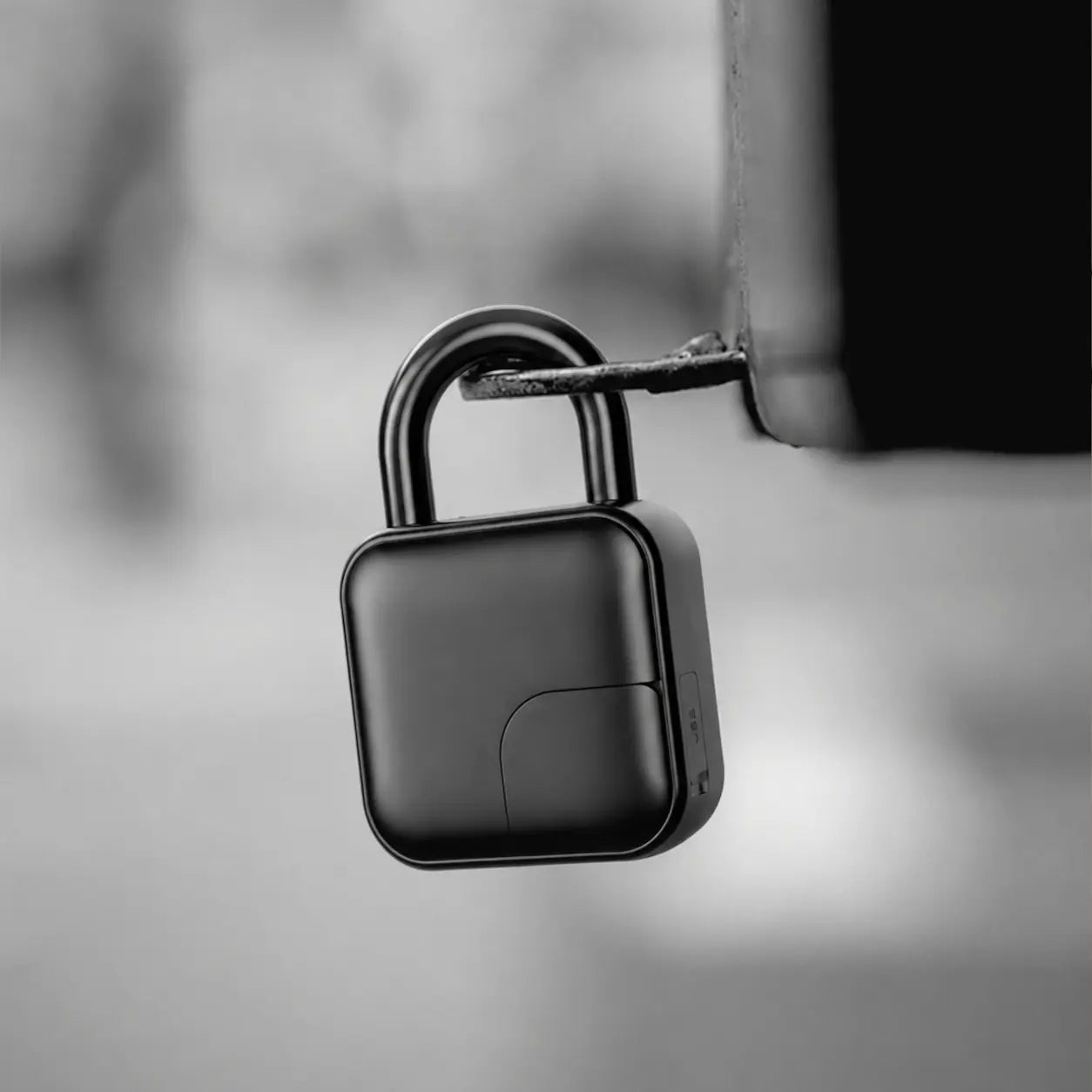 A black square-shaped electronic lock or padlock with a curved shackle, shown hanging from a metal bracket against a blurred gray background.